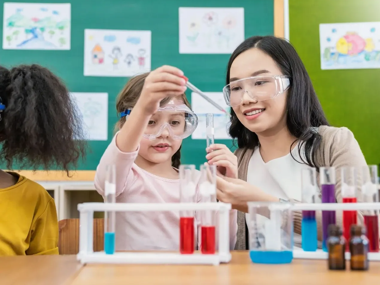  A child wearing safety goggles takes part in a primary school science lesson, assisted by a teacher.