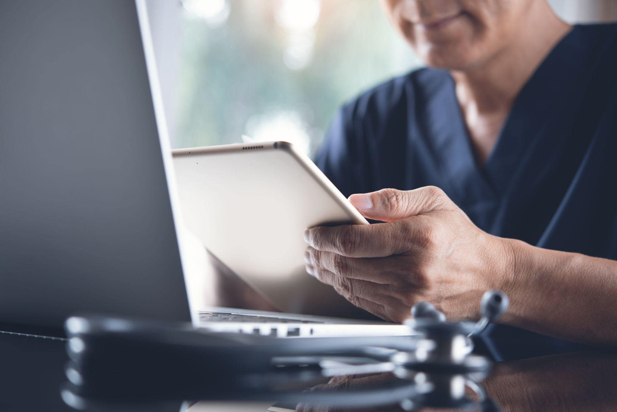 A doctor reviewing a medical record on a tablet with a laptop in front of him