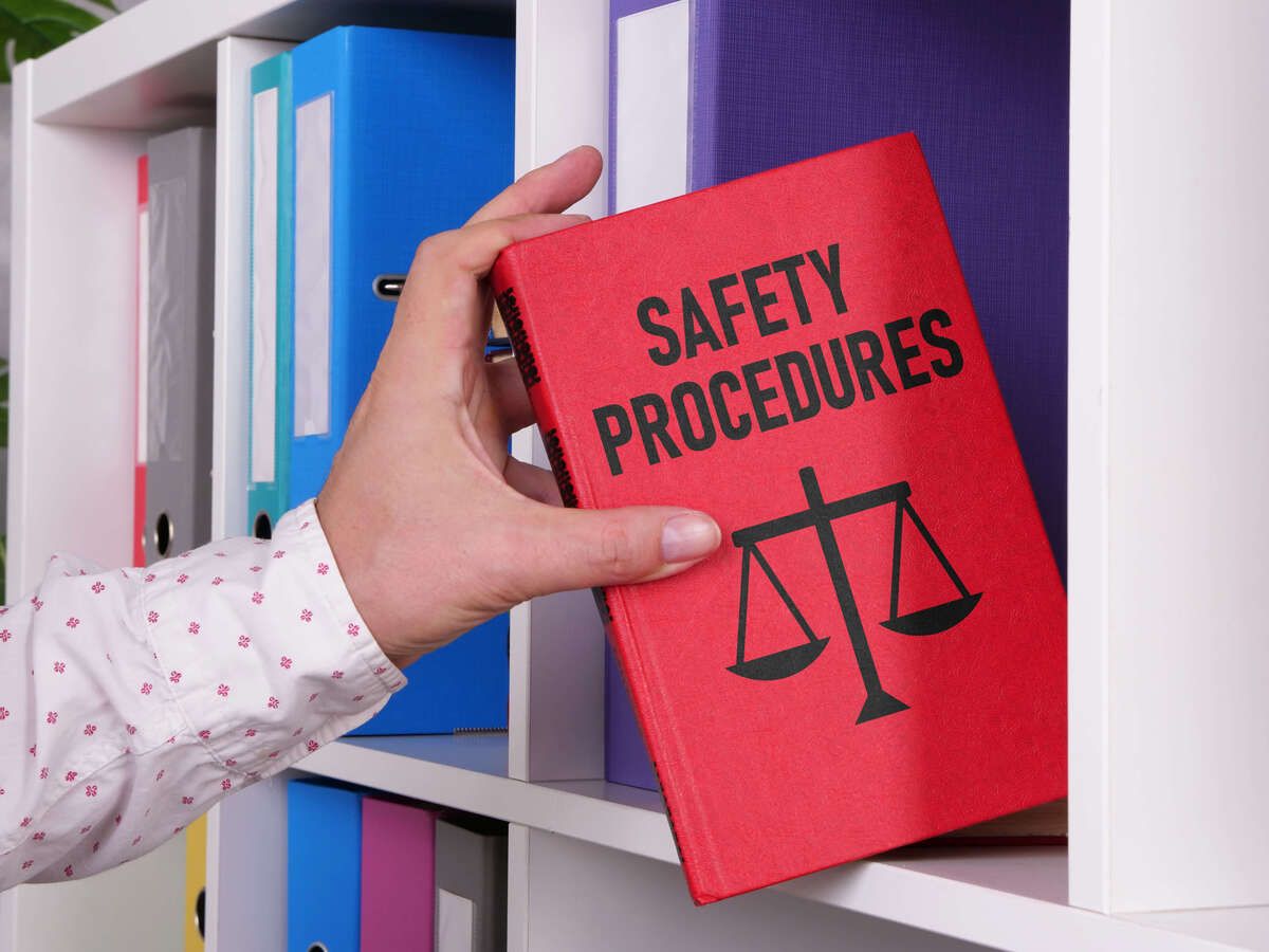 A hand places a red accident book onto a shelf in a school office. The book is titled ‘Safety Procedures’.